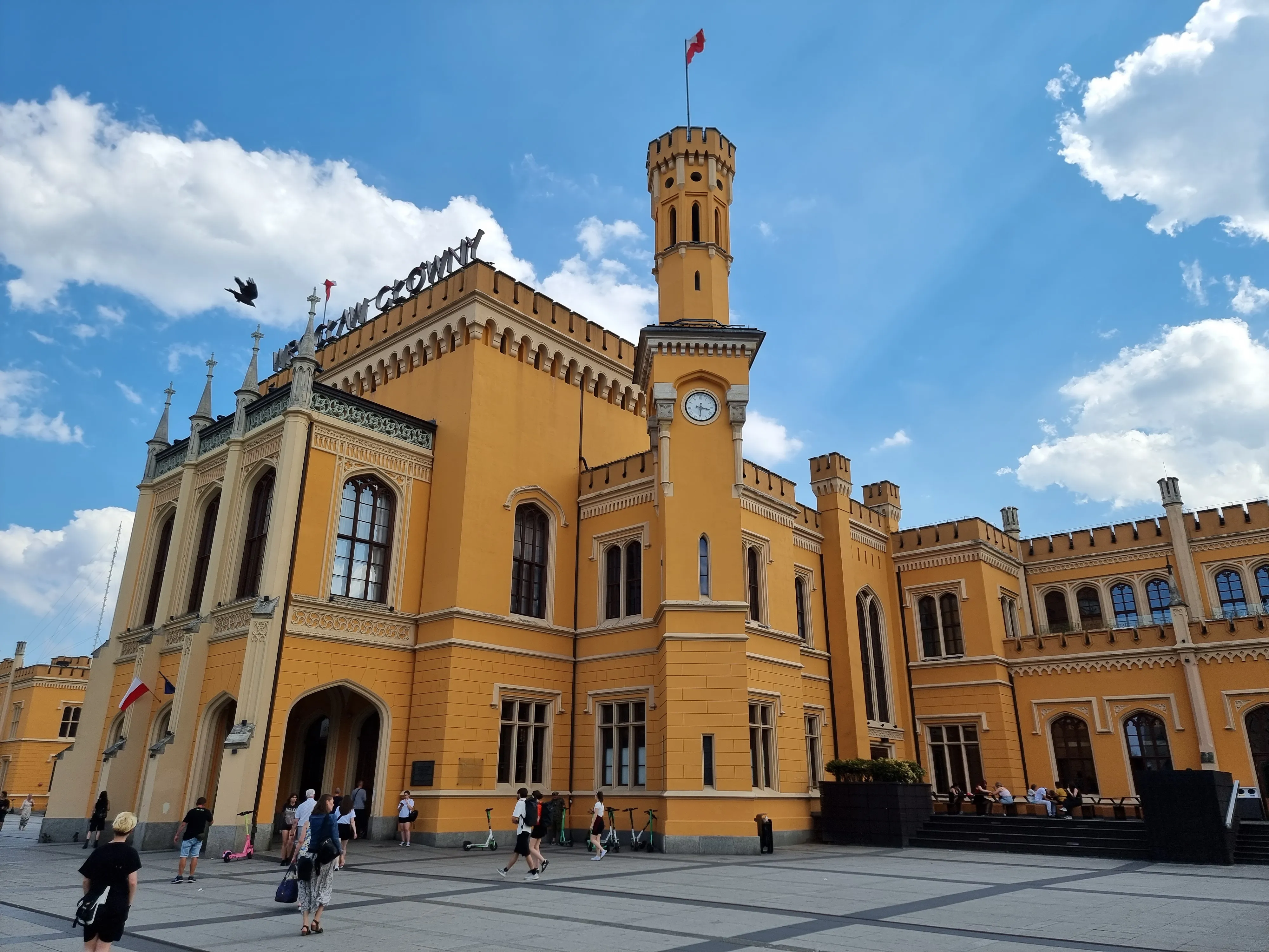 Odnowiona żółta fasada Dworca Głównego z wieżą zegarową i pieszym placem przed wejściem.