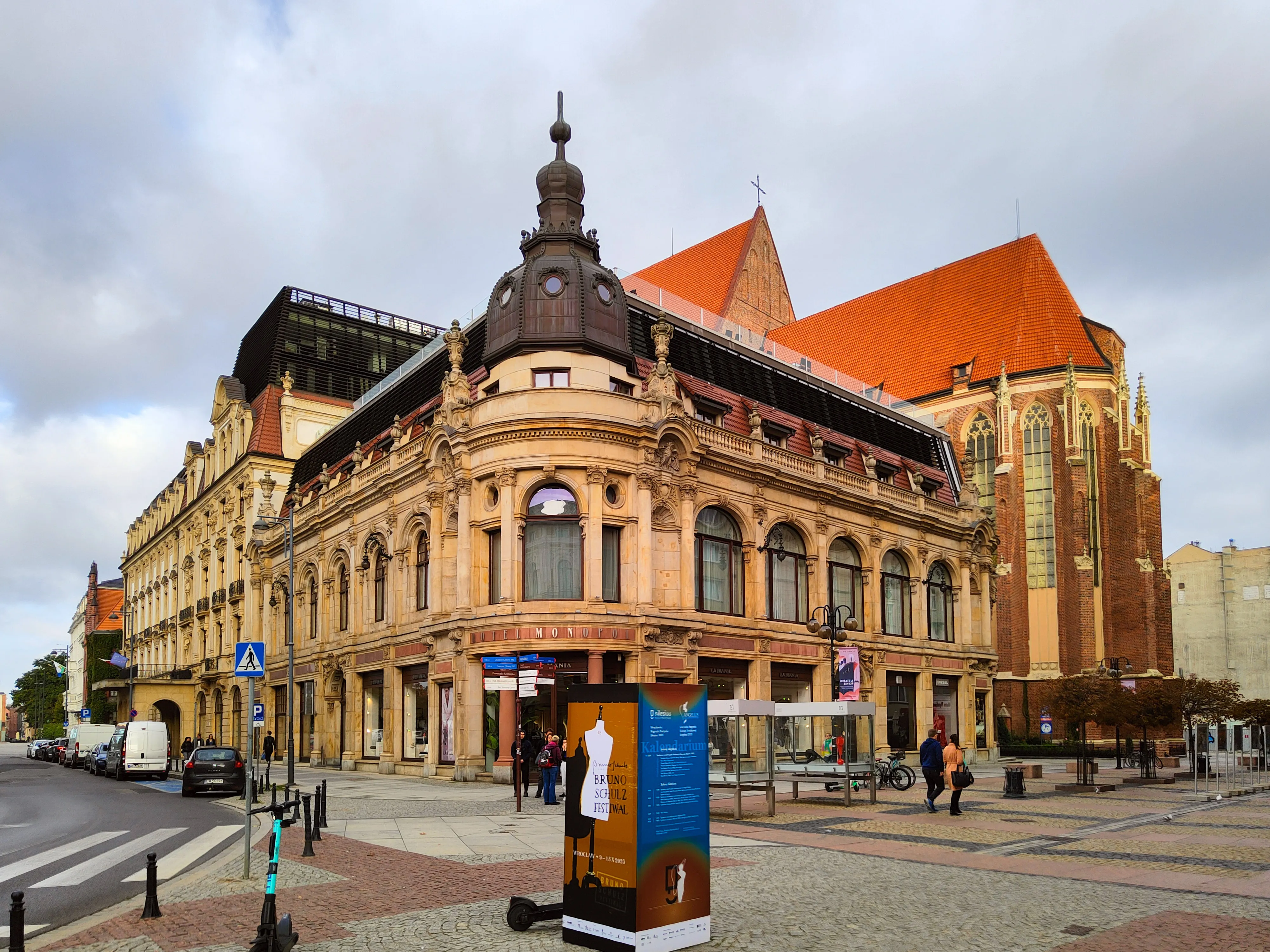 Ten sam narożnik Hotelu Monopol w 2023 roku: odnowiona jasna elewacja, narożna wieżyczka i współczesna nadbudowa; w tle kościół św. Doroty.